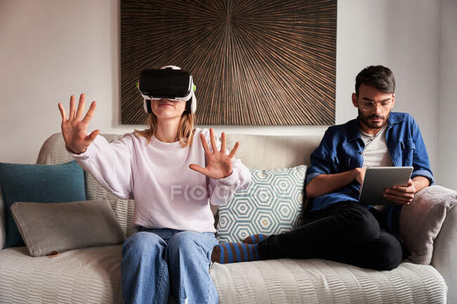 Positive young woman experiencing virtual reality in modern headset sitting on comfortable sofa near concentrated ethnic boyfriend browsing tablet — Stock Photo