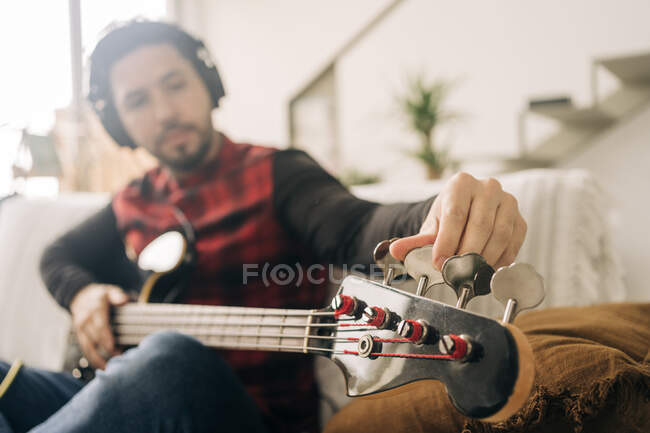 Бородатый мужчина-музыкант в наушниках настраивает бас-гитару, сидя на диване в гостиной — стоковое фото