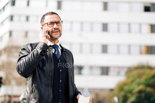 Зрелый латиноамериканец мужчина в элегантной повседневной одежде с горячим напитком, чтобы поговорить по сотовому телефону во время прогулки по городской тротуару — стоковое фото
