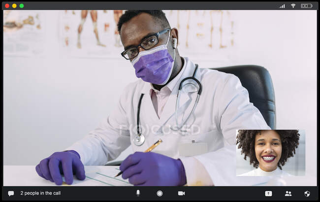 Concentrated African American male doctor in medical robe and mask looking at camera while communicating with positive female during video call — Stock Photo