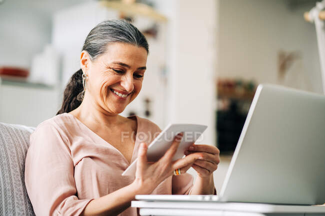Allegra donna di mezza età seduta sul divano vicino al netbook moderno mentre guarda la foto in appartamento moderno con cucina su sfondo sfocato — Foto stock