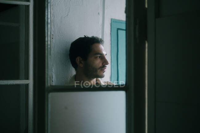 Calma hombre sin camisa con la barba apoyada en la pared en mal estado en casa y mirando hacia otro lado - foto de stock