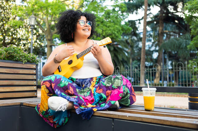 Cheerful female artist in ornamental clothes playing musical instrument on city bench with refreshing drink while looking away — Stock Photo