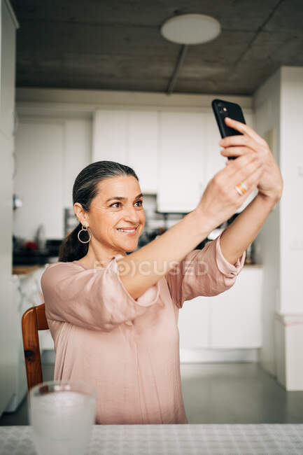 Mulher de meia-idade positiva tomando auto-retrato enquanto sentado à mesa com vidro de água na cozinha moderna em casa — Fotografia de Stock