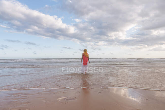 Вид ззаду анонімної жінки, що йде у хвилястій воді величезного океану на піщаному пляжі під хмарним небом — стокове фото