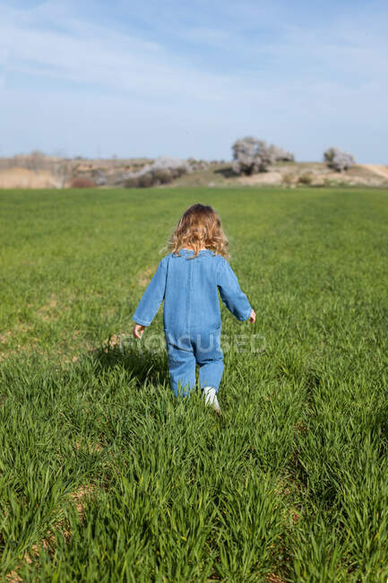 Visão traseira de corpo inteiro da menina irreconhecível em roupas elegantes e boné em pé na grama no dia ensolarado de verão no campo — Fotografia de Stock