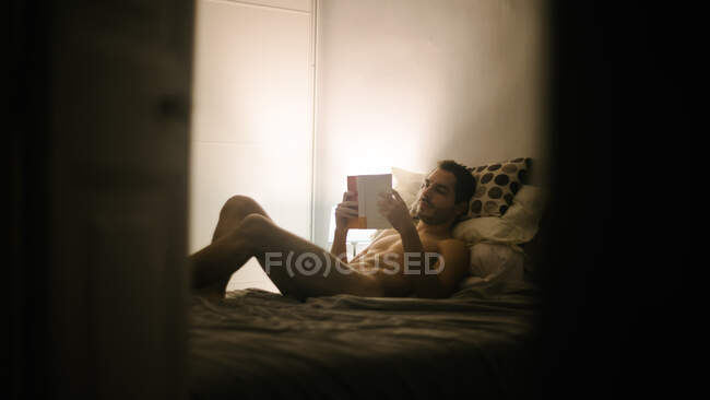 Nu masculino relaxante no confortável cama e leitura interessante livro no quarto no noite — Fotografia de Stock