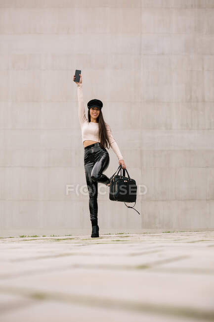 Ground level of delighted female entrepreneur in trendy clothes and with smartphone standing on city street and looking at camera — Stock Photo