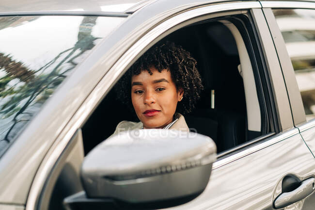Vue latérale de la conductrice afro-américaine sérieuse en tenue à la mode conduisant une automobile moderne dans la rue regardant la caméra — Photo de stock
