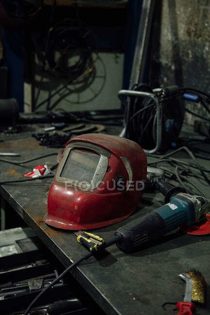 Металлический стол со сварочной маской и угловым шлифовальным станком возле канцелярского ножа и катушки с электрической проволокой в профессиональном гараже — стоковое фото