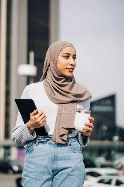 Hembra musulmana en pañuelo para la cabeza caminando con bebida para llevar y tableta en la ciudad mientras mira hacia otro lado - foto de stock