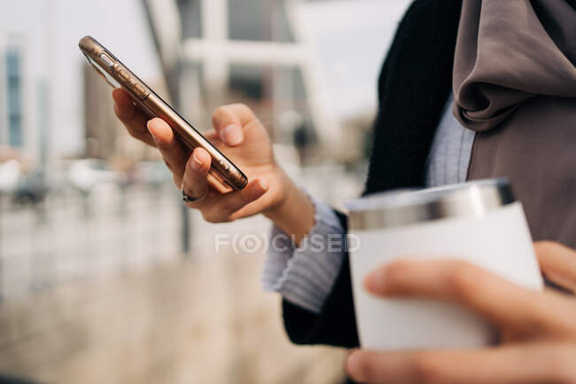 Anonymous ethnic female standing with takeaway drink while surfing Internet on smartphone and enjoying weekend in city — Stock Photo