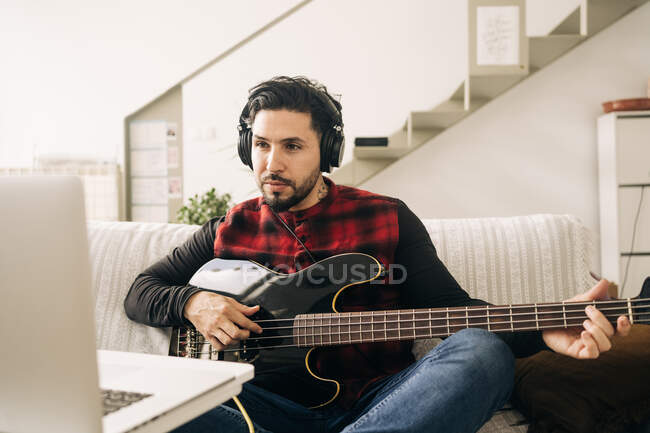 Musicista adulto in cuffia suonare il basso contro netbook sul divano in soggiorno — Foto stock