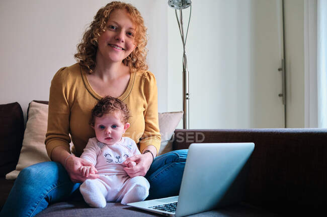 Весела самотня мати-фрилансер сидить з дитиною на дивані і переглядає ноутбук удома вдень. — стокове фото