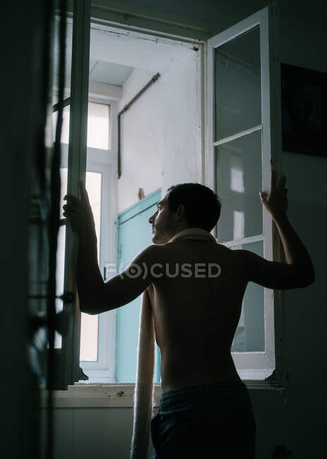 Вид ззаду чоловіка з голим торсом, що стоїть біля вікна вдома і дивиться в сторону — стокове фото