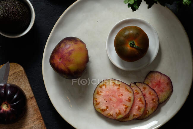 Top view of fresh whole and sliced black tomatoes on table with avocado and green mint during healthy meal preparation — Stock Photo