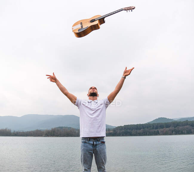 Bearded man in casual clothes standing near lake and tossing guitar in air against hills under gray cloudy sky — Stock Photo