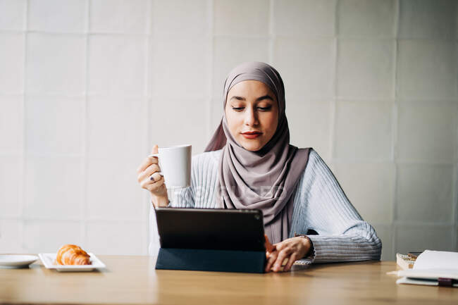 Libero professionista musulmano pensieroso in velo seduto a tavola con tazza di bevanda e tablet mentre pensa al progetto e guardando lo schermo — Foto stock