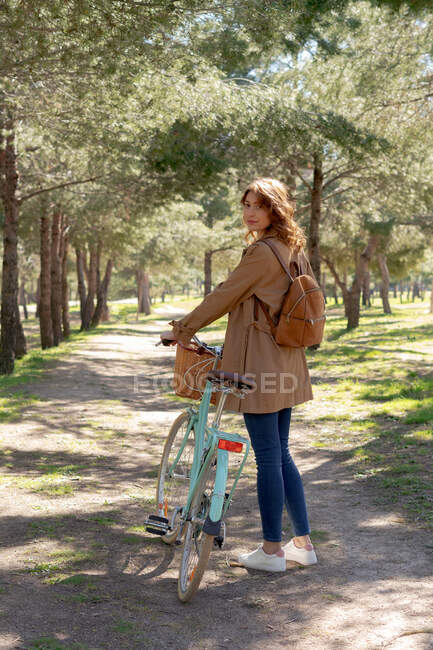Full body of young female standing and concentrated near old bicycle with timber wicker basket in the park while looking at camera — Stock Photo