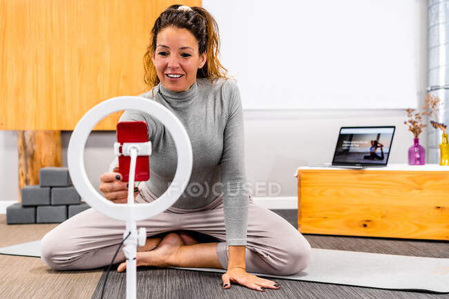 Corps complet de femme athlétique adulte positive en vêtements de sport mettre en place smartphone sur anneau selfie tout en étant assis sur le sol avant de filmer vlog pendant l'entraînement de yoga — Photo de stock