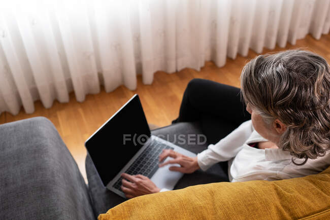 Vista superior de la trabajadora anónima a distancia navegando por Internet en netbook en el sofá en casa - foto de stock