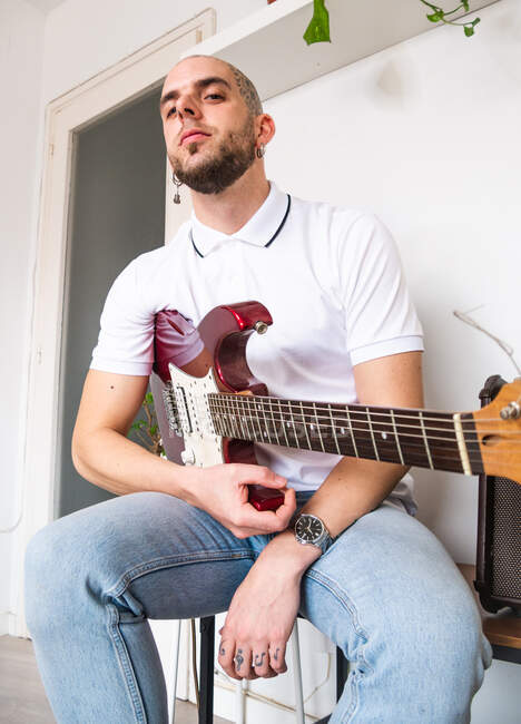 Снизу сконцентрированный музыкант в повседневной одежде сидит с электрогитарой в светлом помещении с белыми стенами в дневное время — стоковое фото