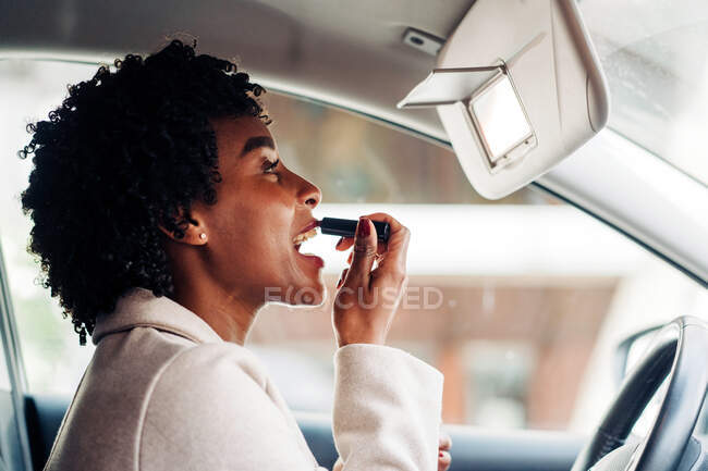 Vue latérale de la conductrice afro-américaine assise dans l'automobile et appliquant du rouge à lèvres tout en regardant le miroir — Photo de stock