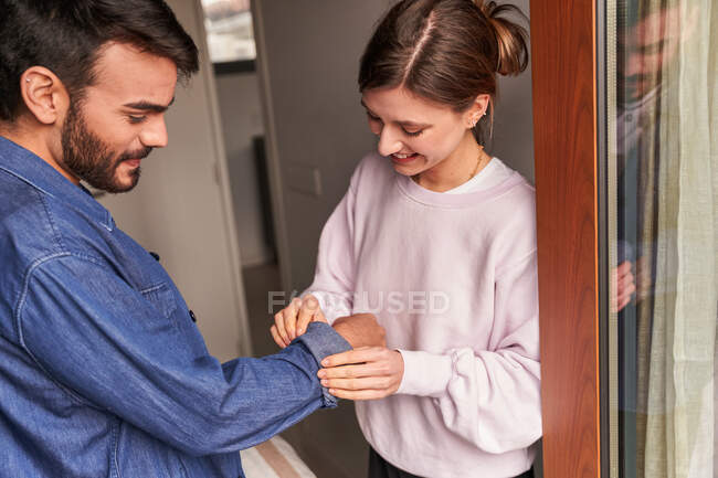 Cortar feliz jovem fêmea arregaçando mangas de camisa jeans de namorado hispânico barbudo enquanto estão juntos perto da janela em casa — Fotografia de Stock