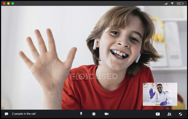 Delighted boy in casual clothes and true wireless earphones smiling and waving hand while talking to positive African American male physician during video conference — Stock Photo