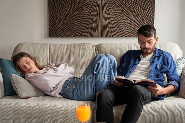 Втомлена молода жінка в повсякденному одязі спить на зручному дивані біля зосередженого етнічного хлопця читання книги в денний час — стокове фото