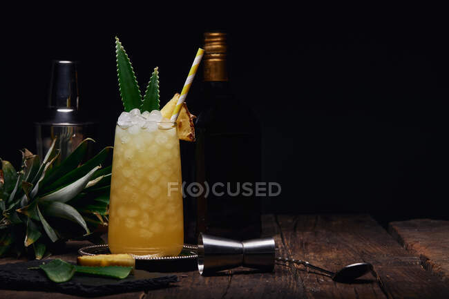Glass of alcoholic cocktail decorated with pineapple piece and leaves with paper straw on tray near shaker and bottle with shot glass at table on black background — Stock Photo