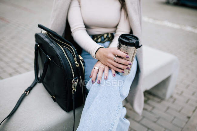 Обрізана невпізнавана жінка-підприємець в модному вбранні, сидячи на лавці з чашкою напоїв, щоб піти — стокове фото