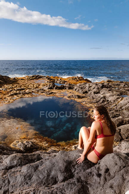 High angle back view of female traveler in bikini sitting on stones against majestic blue foaming sea — Stock Photo