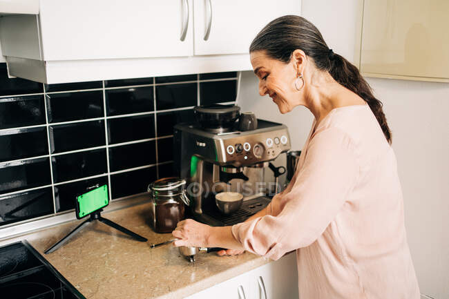 Вид сбоку женщины средних лет с ложкой, наливающей молотый кофе в портафильтр, стоя у кухонной стойки с баночкой кофе и кофемашиной со смартфоном на штативе — стоковое фото