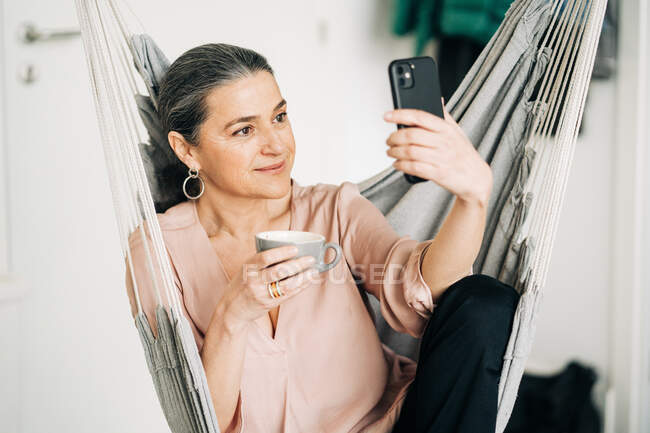 Позитивна жінка середнього віку з чашкою гарячого напою, що має відеочат на смартфоні, сидячи в зручному гамаку вдома на розмитому фоні — стокове фото