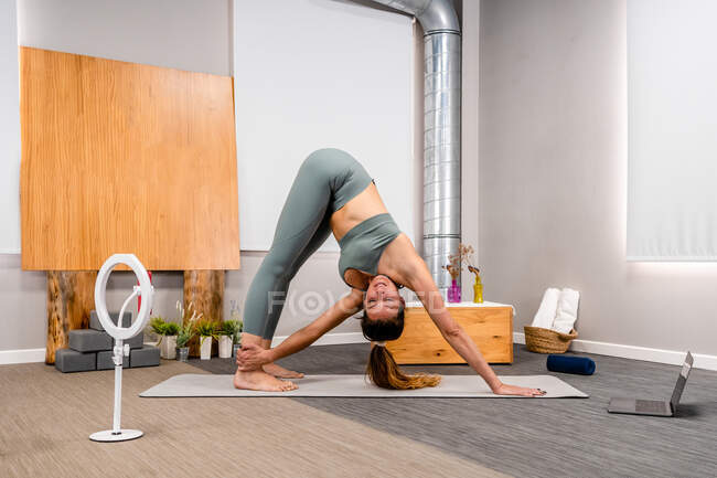 Side view of flexible female in sportswear shooting vlog while performing Revolved Downward Facing Dog asana during yoga session in modern apartment — Stock Photo