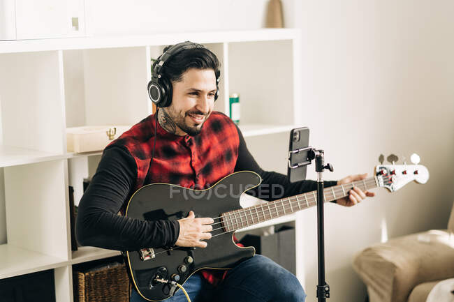 Trípode con cámara colocada cerca del hombre tocando la guitarra en la sala de casa - foto de stock