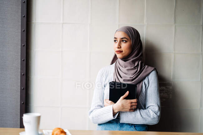 Мусульманська жінка-фрилансер, одягнена в традиційний хіджаб, стоїть у кафе з табличкою під час роботи на проекті віддалено і озирається геть. — стокове фото