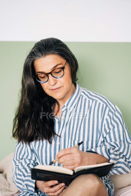 Веселая женщина средних лет в полосатой рубашке улыбается и пишет блокнот на кровати со смартфоном — стоковое фото