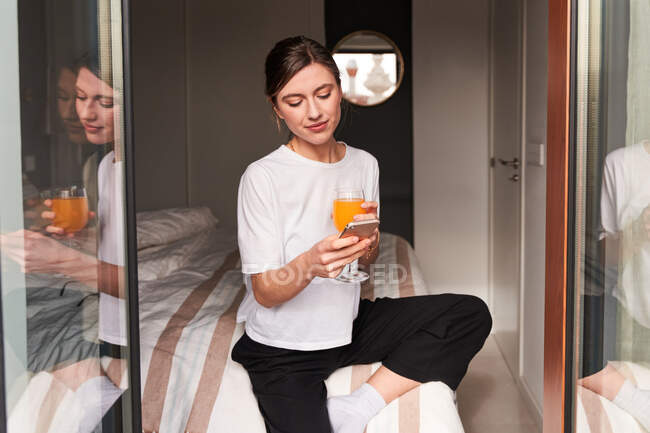 Zufriedene junge Dame in lässiger Kleidung trinkt frischen Orangensaft und Nachrichten auf dem Smartphone, während sie bei Tageslicht im Bett liegt — Stockfoto