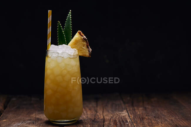 Holztisch mit einem Glas gelbem Cocktail mit Eiswürfeln und erfrischendem Cocktail garniert mit stacheligen Blättern und gestreiftem Stroh — Stockfoto