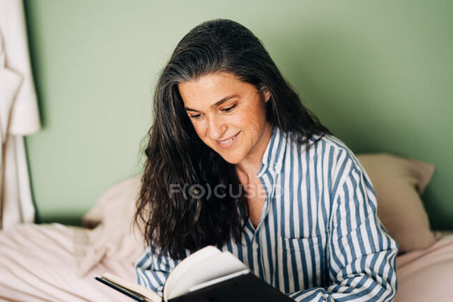 Femme hispanique mature détendue en vêtements décontractés assis au lit et lisant un livre intéressant — Photo de stock