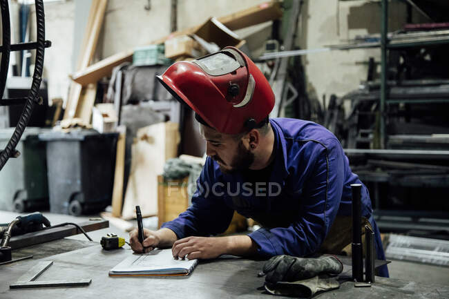 Серйозний бородатий чоловік у хардхаті і взагалі робить нотатки в зошиті з ручкою на столі біля металевих конструкцій у легкому гаражі. — стокове фото