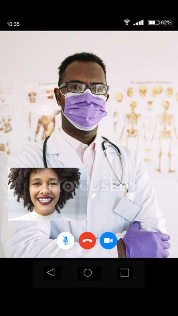 Досвідчений афроамериканський лікар у медичній масці та рукавичках зі стетоскопом, який розмовляє з усміхненою жінкою під час відеобесіди. — стокове фото