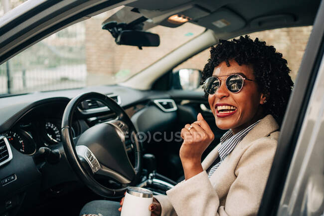 Mulher afro-americana feliz em óculos de sol na moda com caneca térmica sorrindo e deixando automóvel prestígio prata — Fotografia de Stock