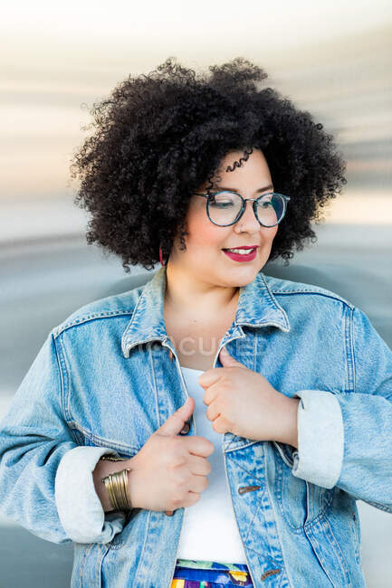 Adult overweight female in trendy clothes and eyewear with Afro hairstyle looking away on blurred background — Stock Photo