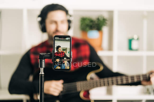 Тренога с экраном мобильного телефона, представляющая фотографию мужчины-музыканта в наушниках, играющего на бас-гитаре в доме — стоковое фото