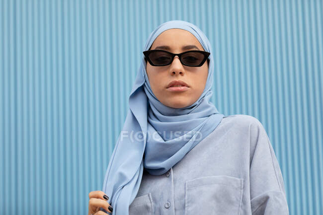 Joven mujer contemplativa étnica en pañuelo azul y gafas de sol modernas mirando a la cámara a la luz del día - foto de stock