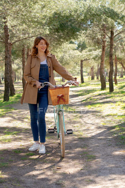 Corpo pieno di allegra giovane femmina sorridente e guardando lontano vicino vecchia bicicletta con cesto di vimini in legno — Foto stock