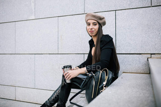 Вид збоку на чудову стильну жінку-підприємця в модному вбранні і березовому капелюсі, що сидить на сходах з напоєм, щоб піти в місто і дивитися в сторону — стокове фото